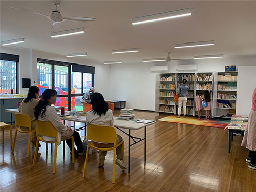 Swanbunko's library room. Three people are sitting at a table in the brightly room, with a bookshelf on the wall side, a child and an adult are selecting books